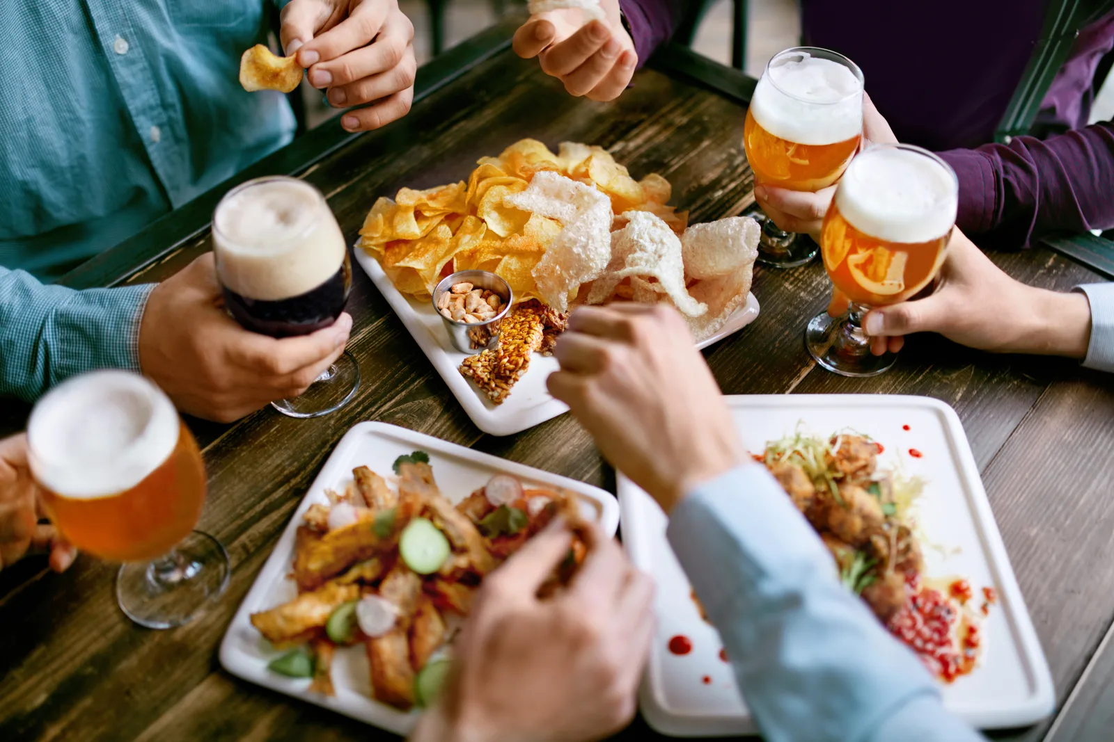 Friends around a dark wooden table sharing wings and crisps, raising glasses of lager and stout.