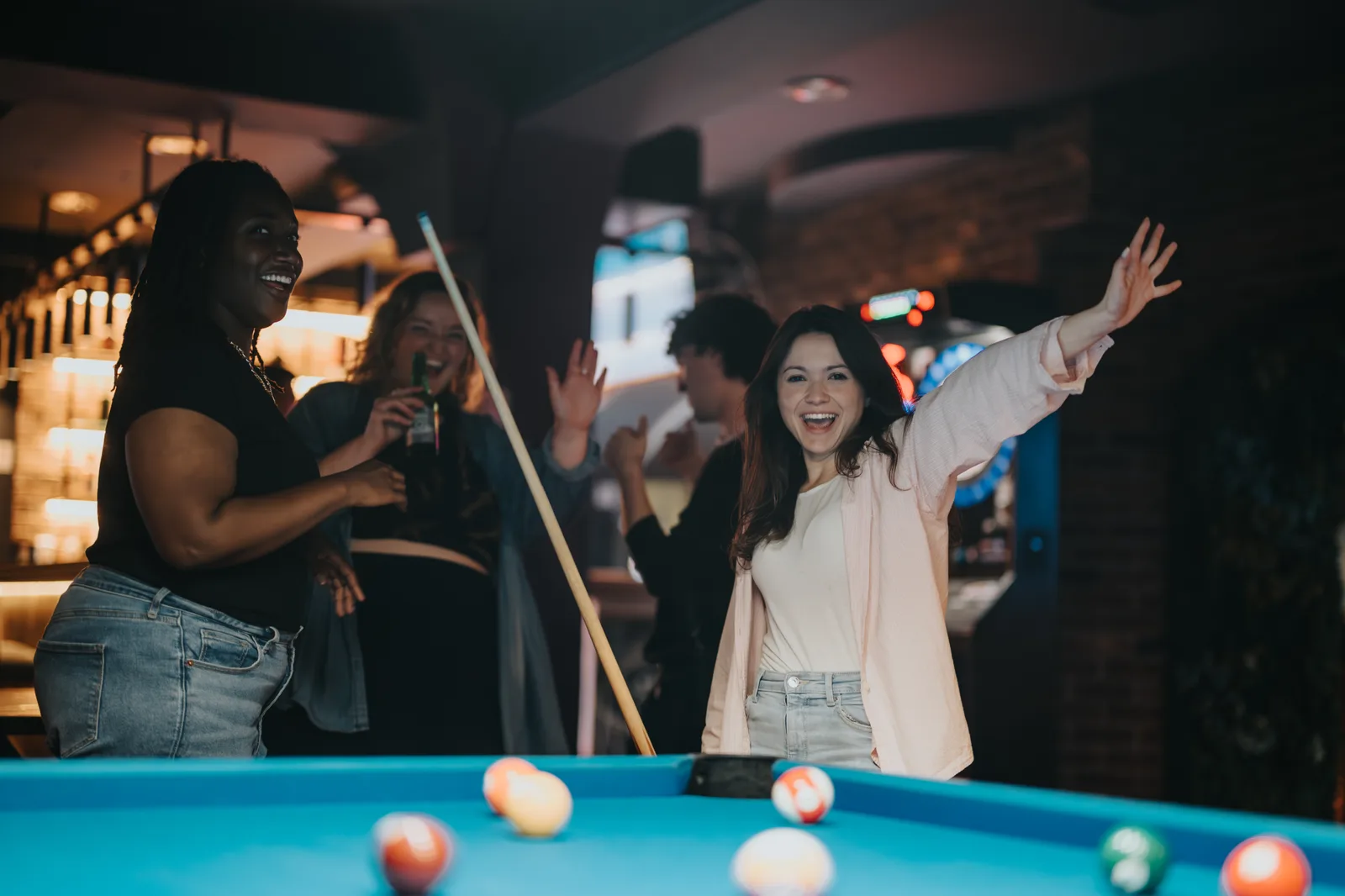 Players laughing around a blue-cloth pool table, one throwing her arms up after a shot while a friend lines up the cue.