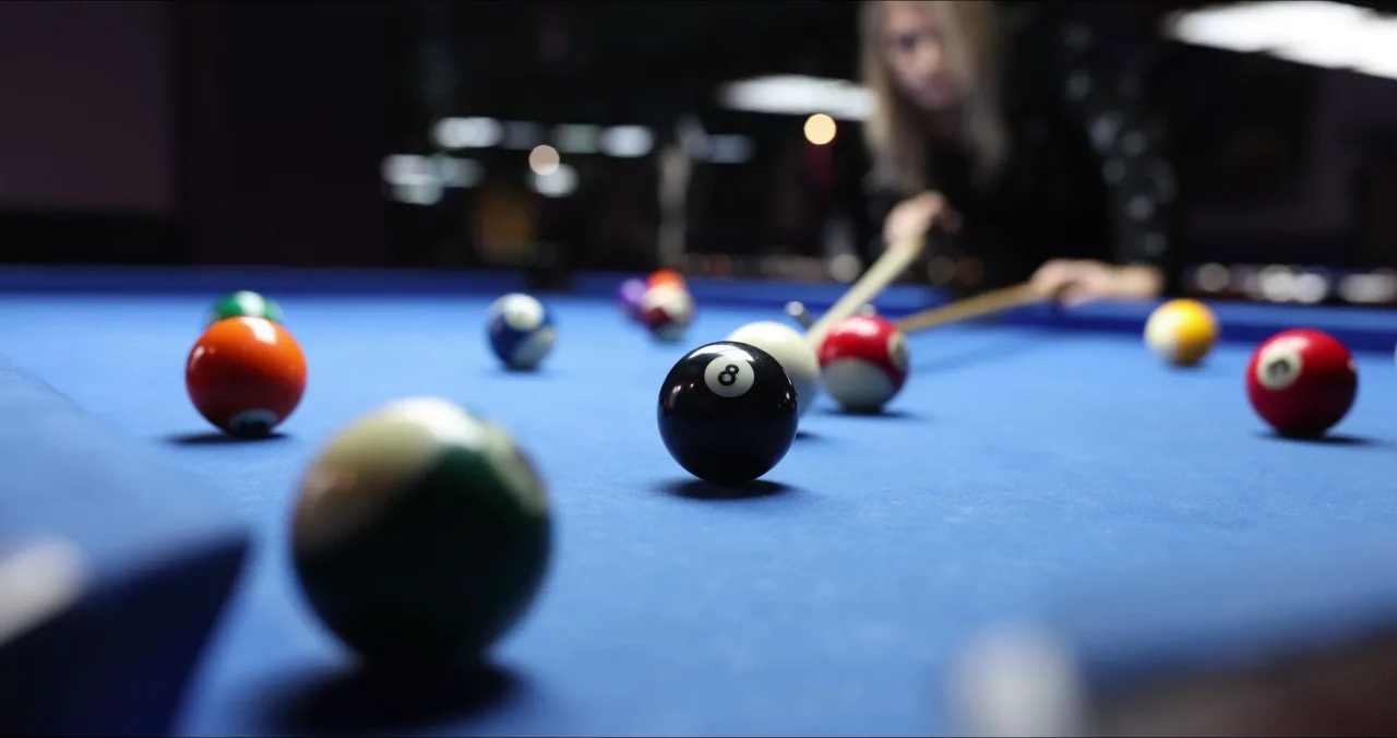 A low view across a blue-cloth pool table with the black 8-ball sharp in the foreground among the scattered colours, and a player leaning in behind it, cue level, lining up her shot in the dark room.