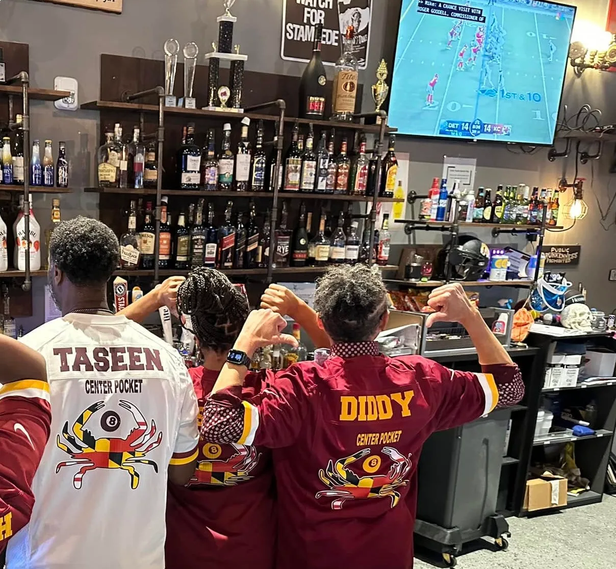 Regulars in team jerseys raising a toast at the bar, football on the screen above the bottle shelves.